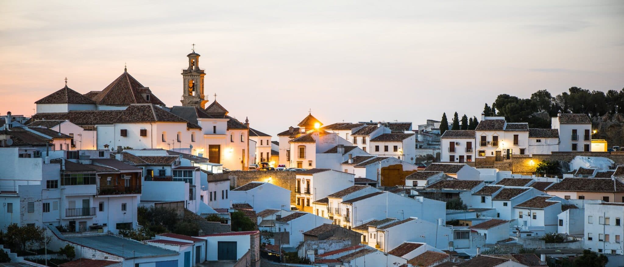 White houses and tiled rooftops of Antequera at dusk, with the church tower lit against a pink sky, Malaga province